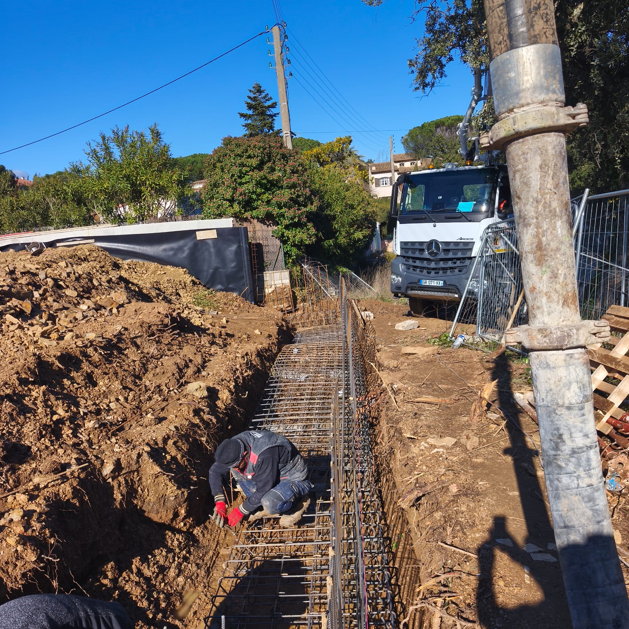 Ouvrier en ferraillage de semelles dans tranchée, camion toupie en arrière-plan, Côte d'Azur